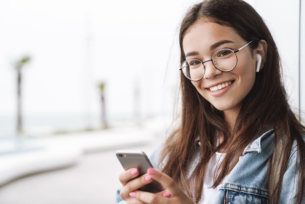 Porträt eines hübschen lächelnden Teenager-Mädchens mit Ohrstöpseln und Brillen mit Smartphone beim Gehen im Freien