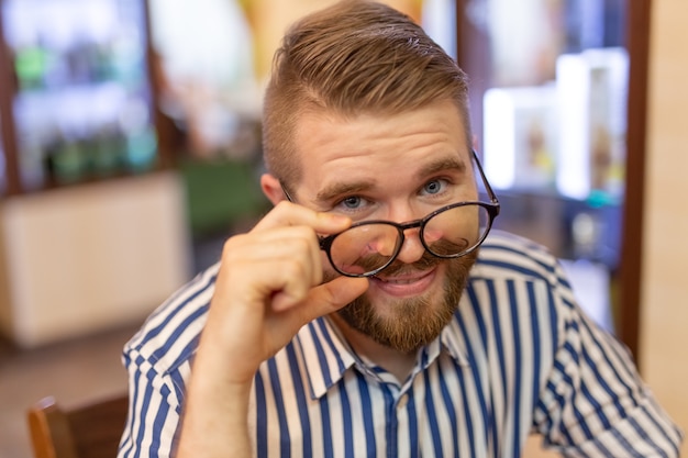 Porträt eines hübschen lustigen jungen Mannes mit einem Schnurrbart und einem Bart, die vorne mit Gläsern in einem Café schauen