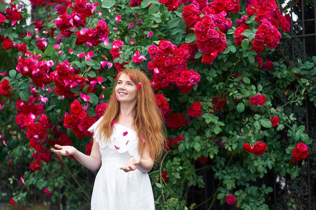 Porträt eines hübschen rothaarigen Mädchens, gekleidet in ein weißes helles Kleid, blühende Rosen. Draussen.