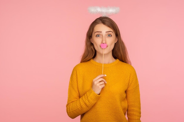 Porträt eines hübschen, schönen Ingwermädchens mit Heiligenschein über dem Kopf, das den Mund mit rosafarbenen Papierlippen auf einem Stock bedeckt, Symbol für Kuss und Liebe, Valentinstag. Studioaufnahme isoliert auf rosa Hintergrund