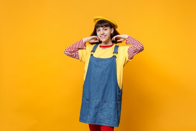 Porträt eines hübschen Teenagers in französischer Baskenmütze, Denim-Sommerkleid, das die Hände in der Nähe von Haaren isoliert auf gelbem Wandhintergrund im Studio hält. Menschen aufrichtige Emotionen, Lifestyle-Konzept. Mock-up-Kopienbereich.