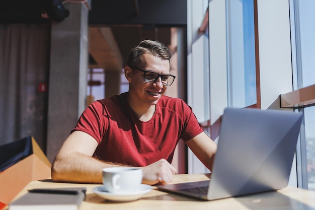 Porträt eines IT-Experten, der remote mit einem modernen Laptop arbeitet, der an einem Tisch sitzt und während einer Pause in die Kamera lächelt, ein glücklicher menschlicher Programmierer mit Sehkorrekturbrille