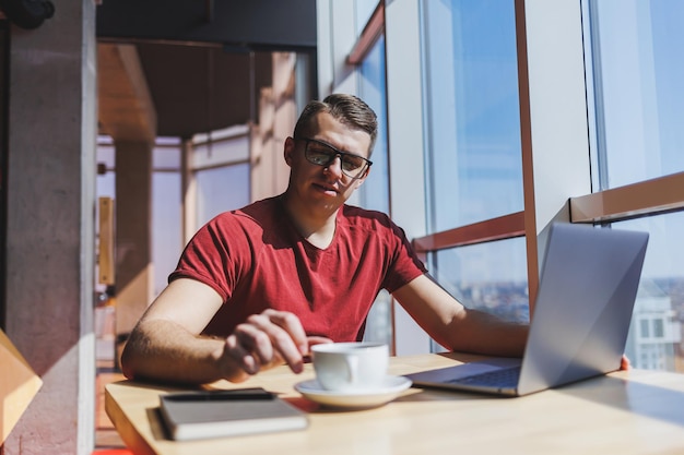 Porträt eines IT-Experten, der remote mit einem modernen Laptop arbeitet, der an einem Tisch sitzt und während einer Pause in die Kamera lächelt, ein glücklicher menschlicher Programmierer mit Sehkorrekturbrille