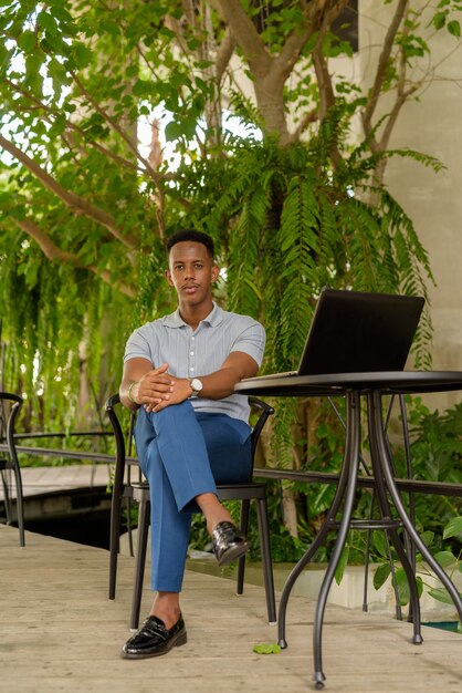 Porträt eines jungen afrikanischen Geschäftsmannes, der Freizeitkleidung trägt und im Café sitzt, während er Laptop-Computer und soziale Distanzierung verwendet