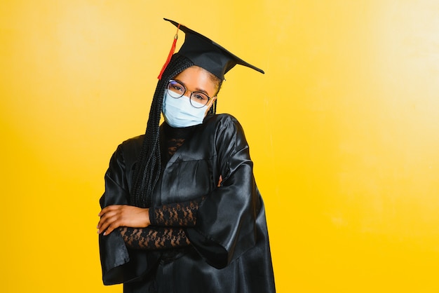 Porträt eines jungen afrikanischen Studenten in einer Graduiertenkappenschutzmaske, auf einer gelben Wand, Graduierung 2021