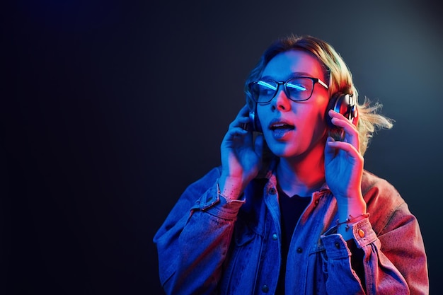 Porträt eines jungen alternativen Mädchens in Brille mit grünen Haaren in rotem und blauem Neonlicht im Studio, das Musik über Kopfhörer hört