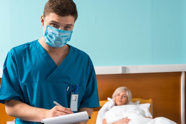 Foto porträt eines jungen arztes in uniform, der eine maske trägt und einige notizen im bericht in der zwischenablage macht, kranker patient liegt im krankenhausbett im hintergrund. gesundheitskonzept