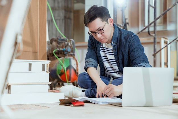 Porträt eines jungen asiatischen Bauunternehmers, der Bauplan auf dem Boden überprüft, mit Konzentration und Fokus zu Hause, um den Baustellenhintergrund zu renovieren