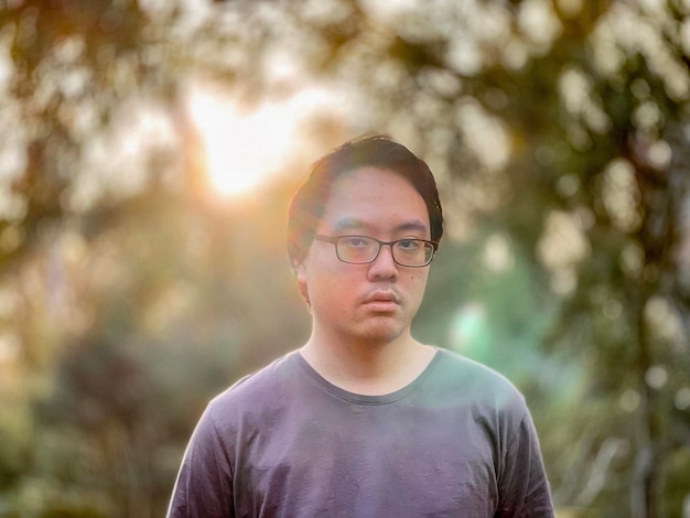 Foto porträt eines jungen asiatischen mannes mit brille, der gegen die untergehende sonne und die bäume steht