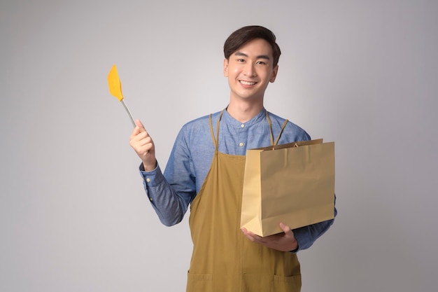 Porträt eines jungen asiatischen Mannes mit Schürze, die Papiertüte und Wender über weißem Hintergrundstudio hält