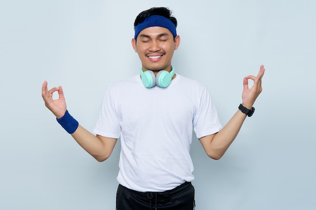 Porträt eines jungen asiatischen sportlichen Mannes mit blauem Stirnband und weißem T-Shirt mit Kopfhörern, die Hände in Yoga-Om-Aum-Geste ausbreiten, entspannen, meditieren, versuchen, sich isoliert auf weißem Hintergrund zu beruhigen