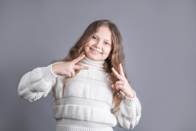 Porträt eines jungen attraktiven kleinen Mädchens mit blondem Haar im Pullover, das V-Zeichen, Friedenszeichen, Siegesgeste auf beiden Händen auf einem grauen Studiohintergrund zeigt.