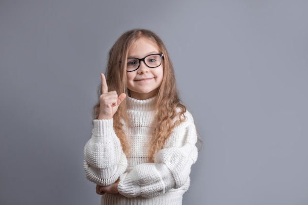 Porträt eines jungen attraktiven kleinen Mädchens mit blondem Haar in einem lächelnden weißen Pullover, der zeigt und Finger oben zeigt, Idee auf einem grauen Studiohintergrund. Platz für Text.