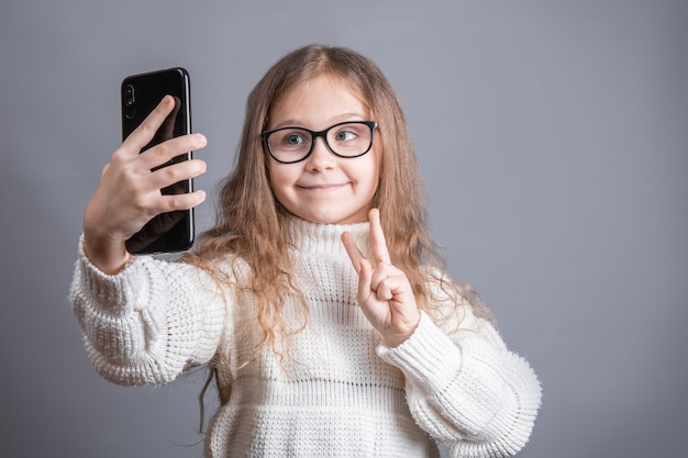 Porträt eines jungen attraktiven kleinen Mädchens mit blonden langen fließenden Haaren in einem weißen Pullover halten Handy nimmt ein Selfie Gespräche über Videokommunikation