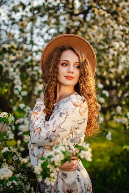 Porträt eines jungen attraktiven Mädchens, das beige Hut und weißes Kleid unter blühenden Bäumen im Park trägt. Frau genießt Sonne und riecht Blumen. Frühlingskonzept.