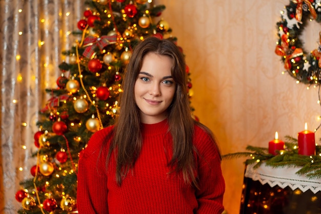 Porträt eines jungen attraktiven Mädchens in rotem Pullover, das auf die Kamera blickt, die in einem geschmückten Raum mit Weihnachtsbaum im Hintergrund steht Weihnachtskonzept feiern