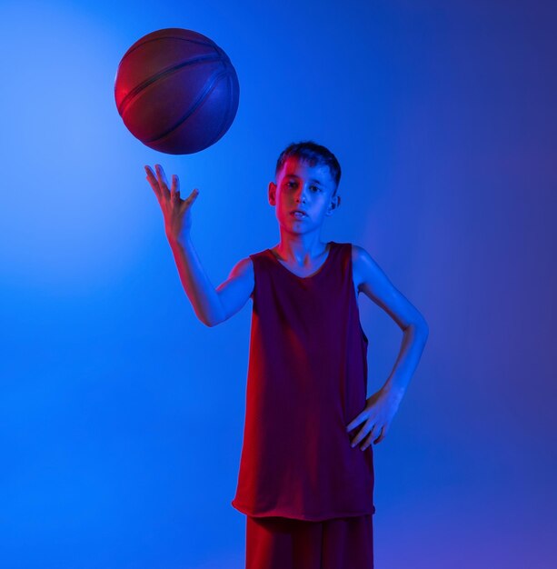 Porträt eines jungen Basketballspielers in Uniform mit Ball, der isoliert über einem blau-violetten Farbverlauf posiert