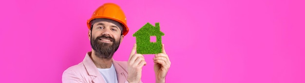 Porträt eines jungen Bauingenieurs mit orangefarbenem Schutzhelm in einer rosafarbenen Jacke, die auf rotem Studiohintergrund steht Ein Mann hält ein grünes Öko-Haus