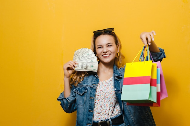 Porträt eines jungen blonden glücklichen Mädchens, das Banknoten und Einkaufstasche über gelber Wand hält