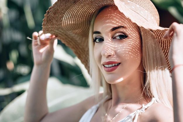 Porträt eines jungen blonden Mädchens mit hellem Make-up im weißen Kleid und Strohhut auf der Straße, das an der Kamera posiert und die Sonne genießt. Nahaufnahme Foto.