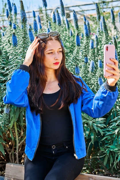 Porträt eines jungen brünetten Mädchens mit gemalten Lippen und in einer blauen Jacke, die ein Selfie macht, das auf der Holzbank im Park sitzt