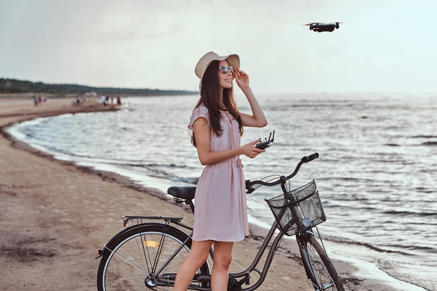 Porträt eines jungen brünetten Mädchens mit Sonnenbrille und Hut im Kleid genießen Sie den Urlaub am Strand und spielen Sie mit dem Quadcopter.