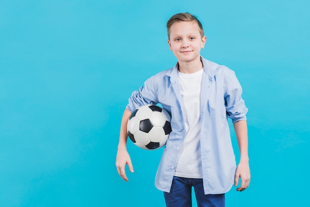 Porträt eines Jungen, der den Fußball schaut zur Kamera steht gegen blauen Himmel hält