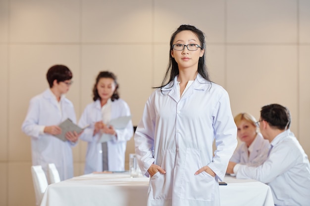 Porträt eines jungen ernsthaften chinesischen Forschers im weißen Laborkittel, der im Konferenzraum steht, ihre Kollegen im Hintergrund sprechen