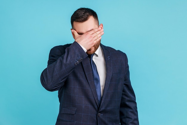 Foto porträt eines jungen erwachsenen im anzug im offiziellen stil, der die augen mit der hand schließt, will das nicht sehen, probleme ignorieren, sich vor stresssituationen verstecken. studioaufnahme im innenbereich isoliert auf blauem hintergrund.