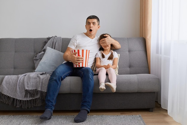 Porträt eines jungen erwachsenen Mannes, der sich zu Hause einen Horrorfilm ansieht, während seine Tochter seinem Kind, das auf dem Sofa sitzt und Popcorn in einem Eimer hält, die Augen bedeckt und den Schock zum Ausdruck bringt, sehr beeindruckt zu sein