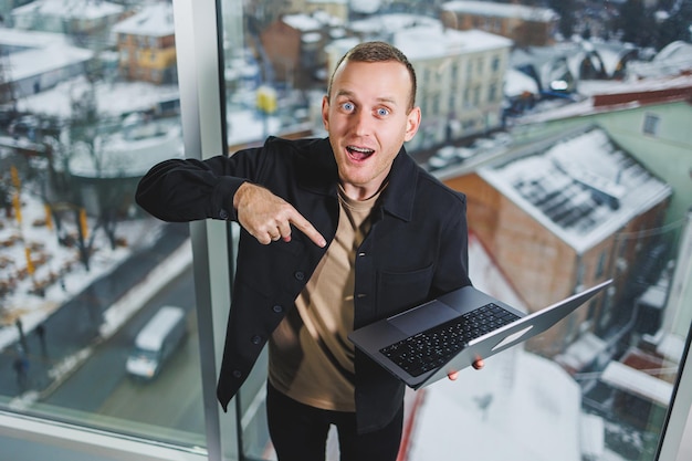 Foto porträt eines jungen fröhlichen mannes mit einem laptop in seinen händen, der nahe dem fenster steht arbeit im büro erfolgreicher männlicher manager