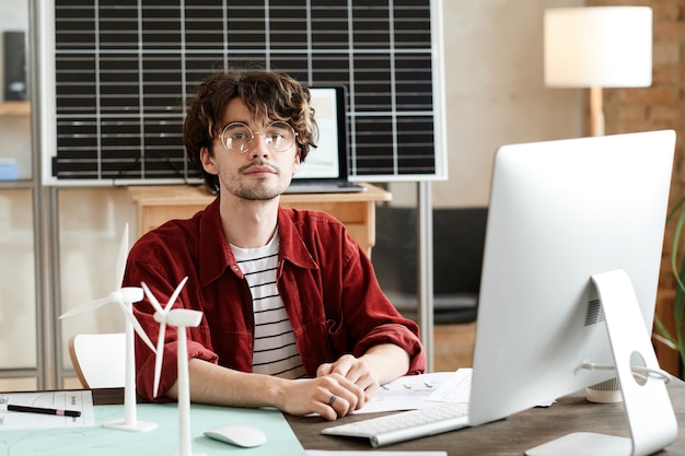 Porträt eines jungen Geschäftsmannes mit Brille, der die Kamera anschaut, während er an seinem Arbeitsplatz mit Computer im Büro arbeitet