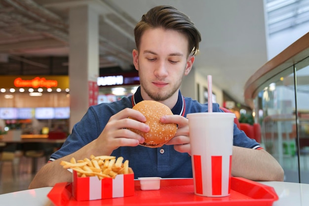 Porträt eines jungen, glücklichen, gutaussehenden Mannes, der hungrig ist, sitzt auf einem Food Court in einem Einkaufszentrum und isst in den Händen einen saftigen, leckeren großen Burger, der Hamburger anschaut Fast Junk ungesundes Fett Essen Hunger