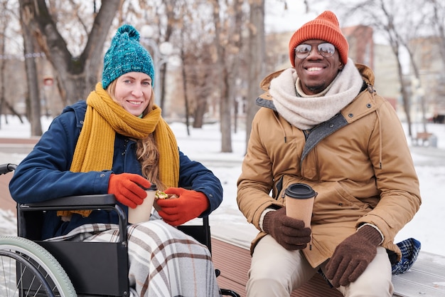 Porträt eines jungen glücklichen Paares, das in die Kamera lächelt, während es im Winterpark sitzt und heißen Kaffee trinkt