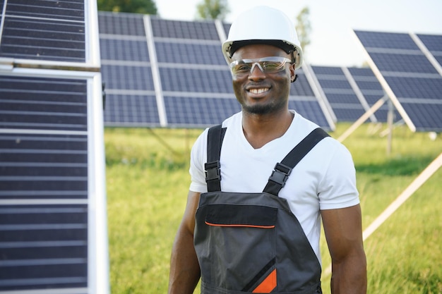 Porträt eines jungen, gutaussehenden afroamerikanischen Handwerkers mit Schutzhelm. Mann in Uniform und mit Werkzeugen, der zwischen Sonnenkollektoren steht