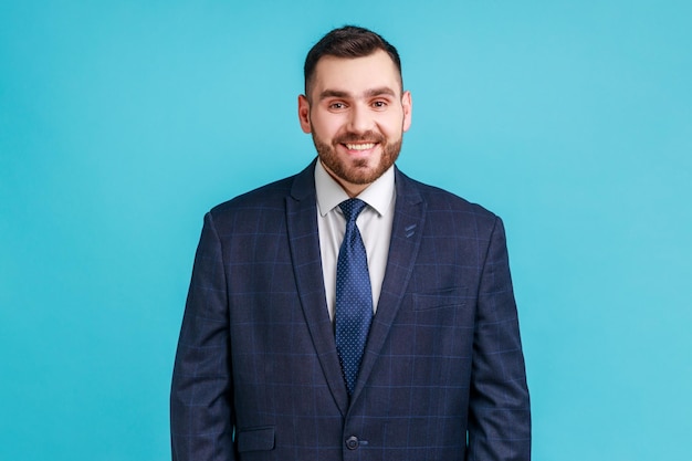 Porträt eines jungen, gutaussehenden Geschäftsmannes, der einen Anzug im offiziellen Stil trägt und mit einem zahnigen Lächeln in die Kamera blickt, das positive Emotionen und Glück ausdrückt Studioaufnahme isoliert auf blauem Hintergrund