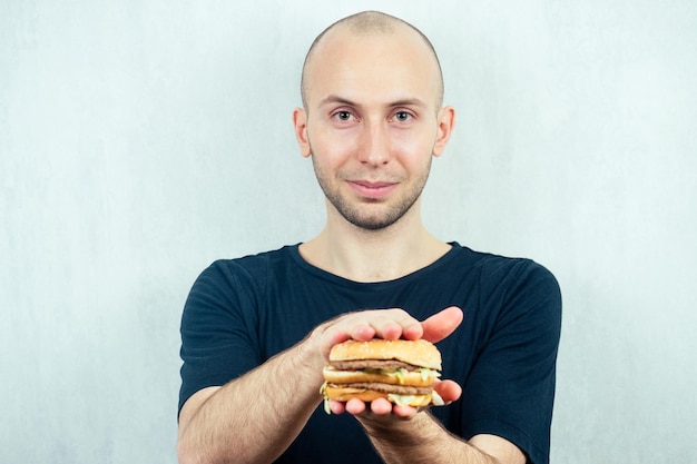 Porträt eines jungen gutaussehenden glatzköpfigen Mannes, der einen kalorienreichen großen Burger in seinen Händen hält. Konzept der Ernährung, ungesunde und gesunde Ernährung