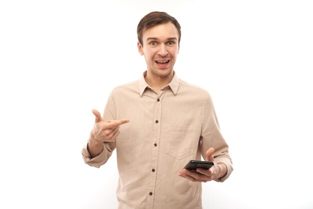 Porträt eines jungen, gutaussehenden kaukasischen Mannes, der ein Mobiltelefon benutzt und lächelnd mit dem Finger auf den Bildschirm zeigt, isoliert auf weißem Studiohintergrund, freudiger Gesichtsausdruck