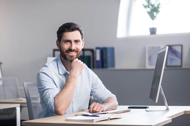 Porträt eines jungen, gutaussehenden Mannes, der an einem Computertisch in einem weißen Büro in einem weißen Hemd sitzt. Er schaut auf die Kamera und lächelt