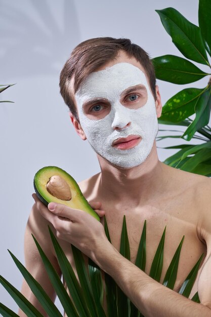 Porträt eines jungen gutaussehenden Mannes mit kosmetischer Maske auf seinem Gesicht, der Avocado in der Hand hält, isoliert auf exotischem Palmblatthintergrund