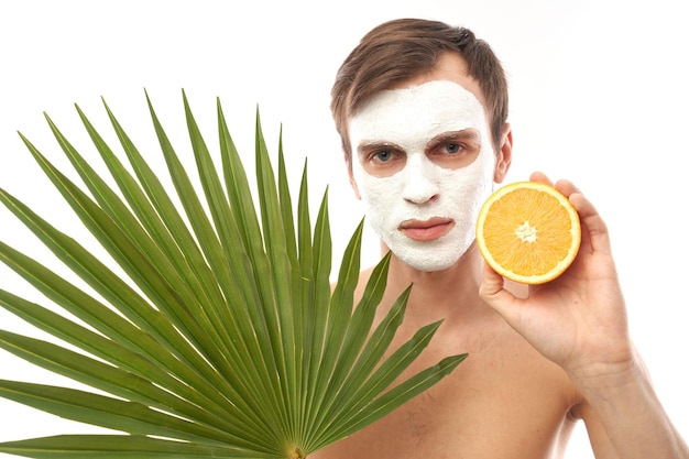 Porträt eines jungen gutaussehenden Mannes mit kosmetischer Maske auf seinem Gesicht, der Orange in der Hand hält, isoliert auf exotischem Palmblatthintergrund