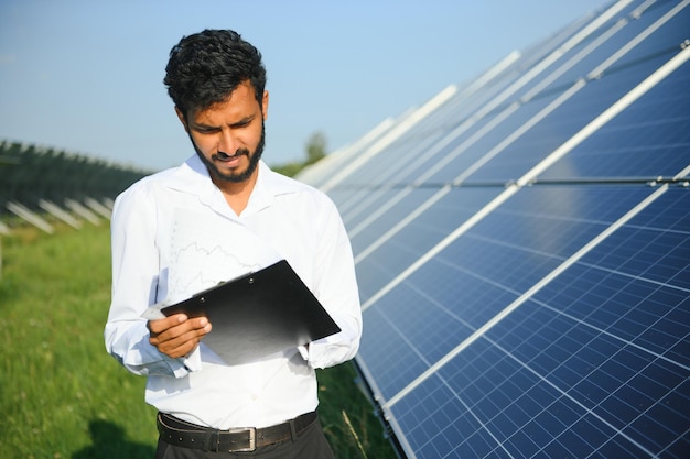 Porträt eines jungen indischen männlichen Ingenieurs, der in der Nähe von Solarpanelen mit klarem blauem Himmel steht.