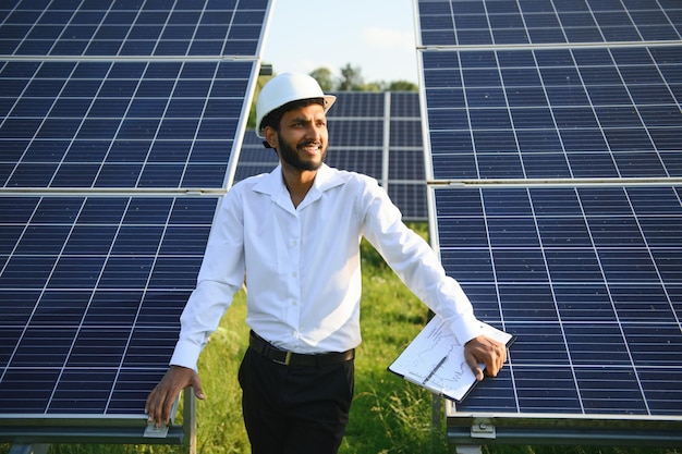 Porträt eines jungen indischen Technikers oder Managers in formeller Kleidung, der vor einem Mann mit Solarpanel für erneuerbare Energien steht und mit verschränkten Armen Kopierraum hat