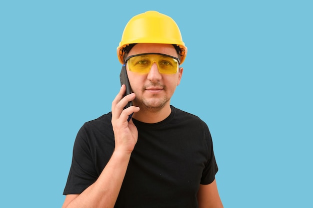 Porträt eines jungen Ingenieurs mit Schutzhelm, der ein Mobiltelefon auf blauem Hintergrund verwendet