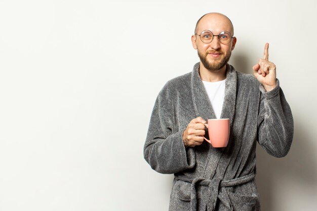 Porträt eines jungen kahlen Mannes mit einem Bart im Bademantel und in den Gläsern, die eine Tasse heißen Kaffee halten