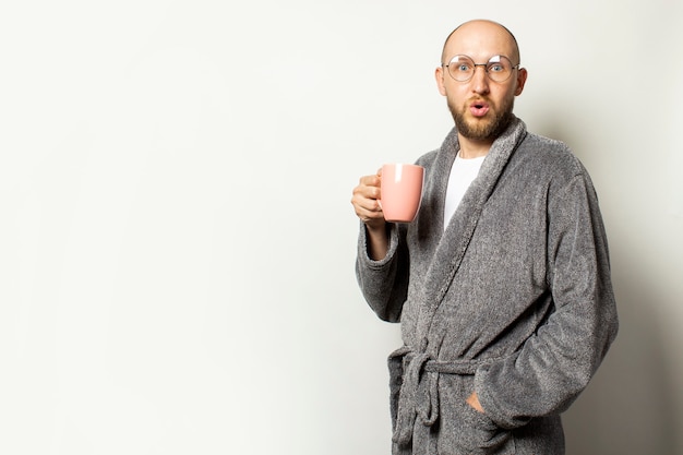 Porträt eines jungen kahlen Mannes mit einem Bart im Bademantel und in den Gläsern, die eine Tasse heißen Kaffee halten