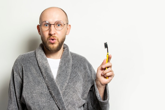 Porträt eines jungen kahlen Mannes mit einem Bart in einem Bademantel und einer Brille und einem überraschten Gesicht und hält eine Zahnbürste in der Hand