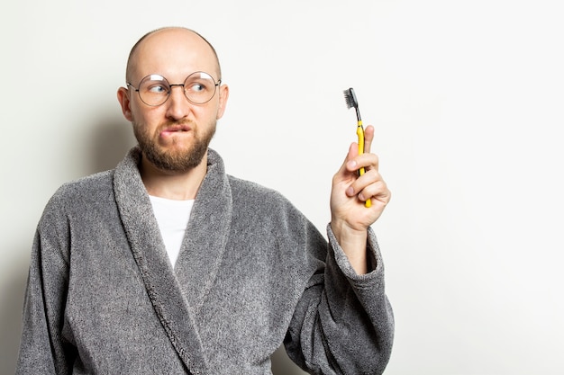 Porträt eines jungen kahlen Mannes mit einem Bart in einem Bademantel und einer Brille und einem überraschten Gesicht und hält eine Zahnbürste in der Hand