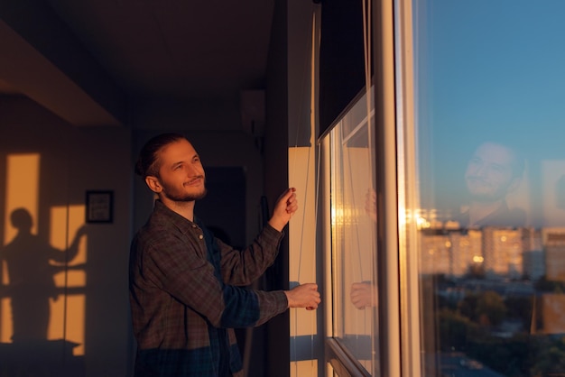 Porträt eines jungen lächelnden Mannes schließt Fenster gerollte Jalousien an einem sonnigen Tag bei Sonnenuntergang