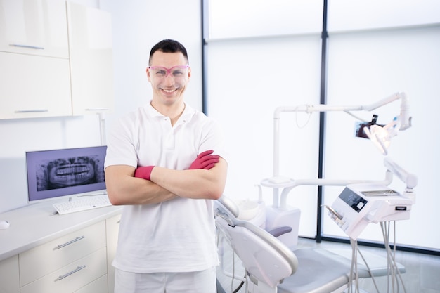 Foto porträt eines jungen lächelnden zahnarztes in der arbeitsbrille und in einer weißen uniform. der mann verschränkte die arme und steht am zahnarztstuhl im büro.